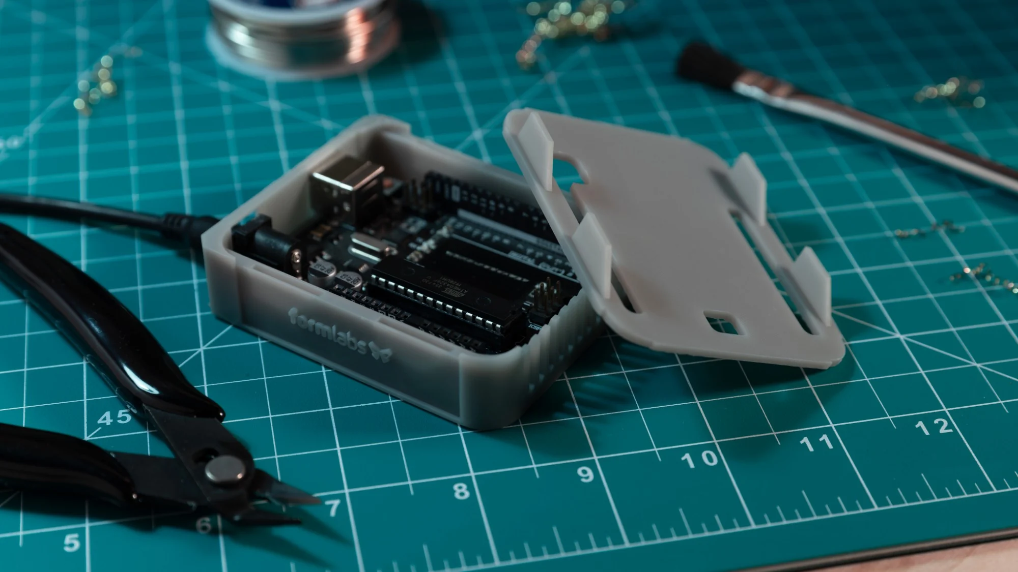 A grey 3D printed Arduino case on a teal cutting mat, an Arduino board inside and its lid with snap-fit clips beside it